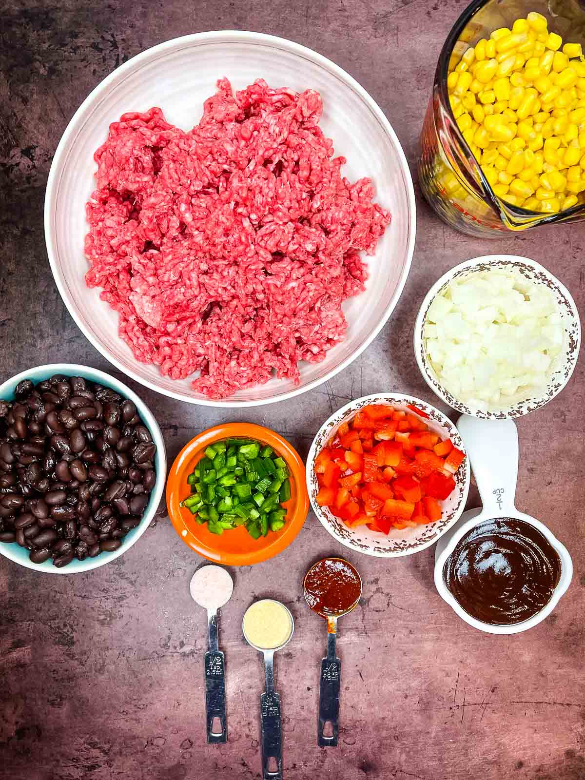 Overhead of ingredients in individual bowls including raw ground beef, corn, black beans, chopped onion, diced red and green peppers seasonings, barbecue sauce and chipotle in adobo.