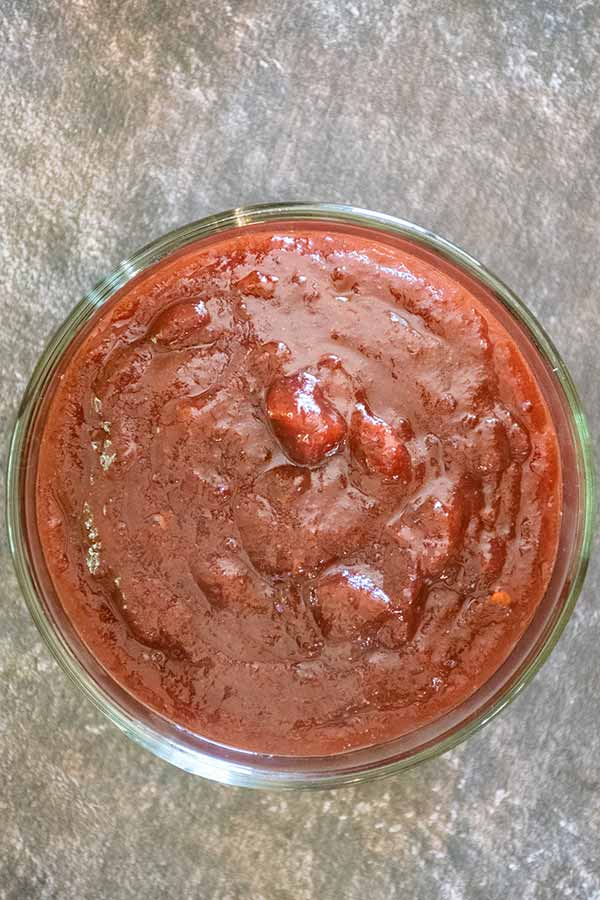 Overhead view of a glass bowl filled with dark red cranberry chipotle sauce on a textured gray surface.