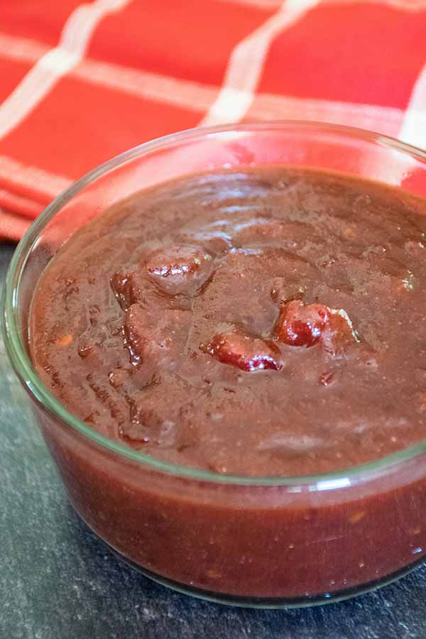 Side angle of a glass bowl filled with dark red textured sauce with a red plaid towel in the background.