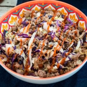 Square image of a colorful bowl filled with ground venison, shredded green and red cabbage, and drizzled with sriracha sauce.