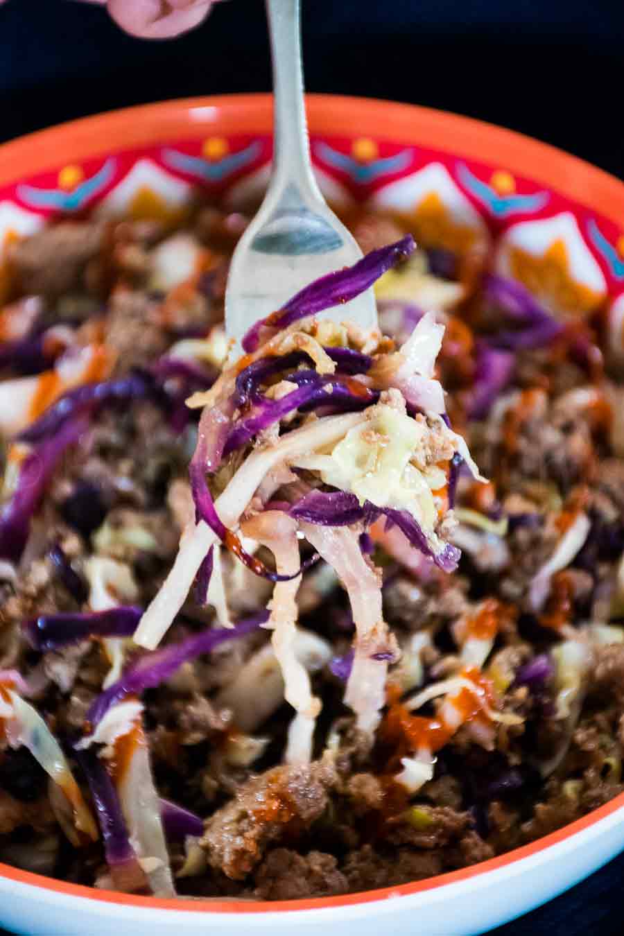 Close up of a forkful of eggroll in a bowl with shredded red and green cabbage, ground meat and a drizzle of sriracha sauce lifted from a colorful bowl.