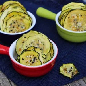 Thin dehydrated zucchini chips seasoned with everything bagel seasoning, served in colorful ramekins on a navy cloth.