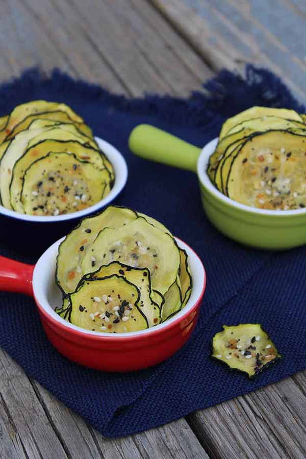 Thin dehydrated zucchini chips seasoned with everything bagel seasoning, served in colorful ramekins on a navy cloth.