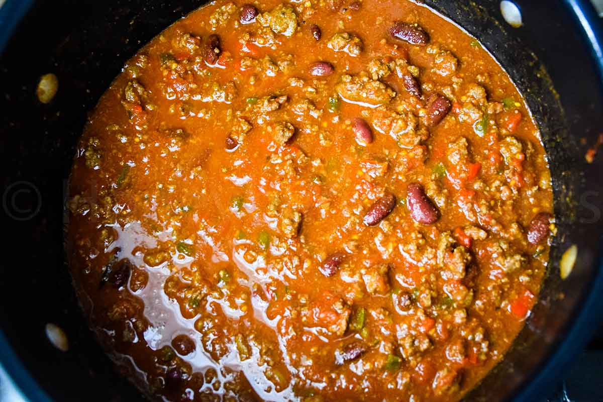 A large pot of simmered chili with visible chunks of ground meat, beans, and peppers in a thick red sauce.