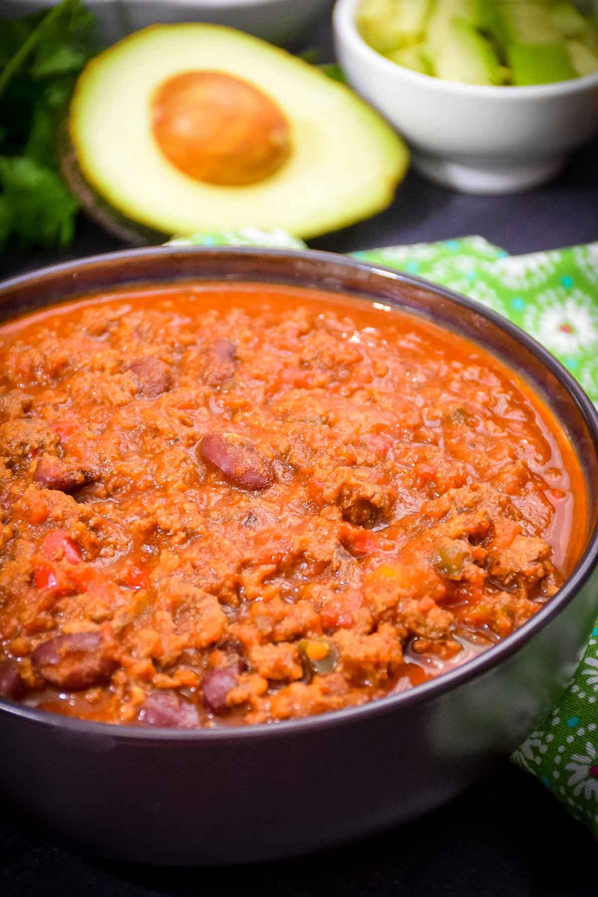 A close up of a bowl filled with chili made with ground venison and beans, with an chopped avocado in a white bowl in the back ground and an avocado half next to it.