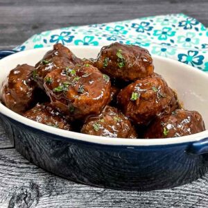 Oval navy blue dish filled with glazed grape jelly BBQ meatballs, garnished with chopped parsley, set on a dark wooden surface with a teal floral napkin in the background.