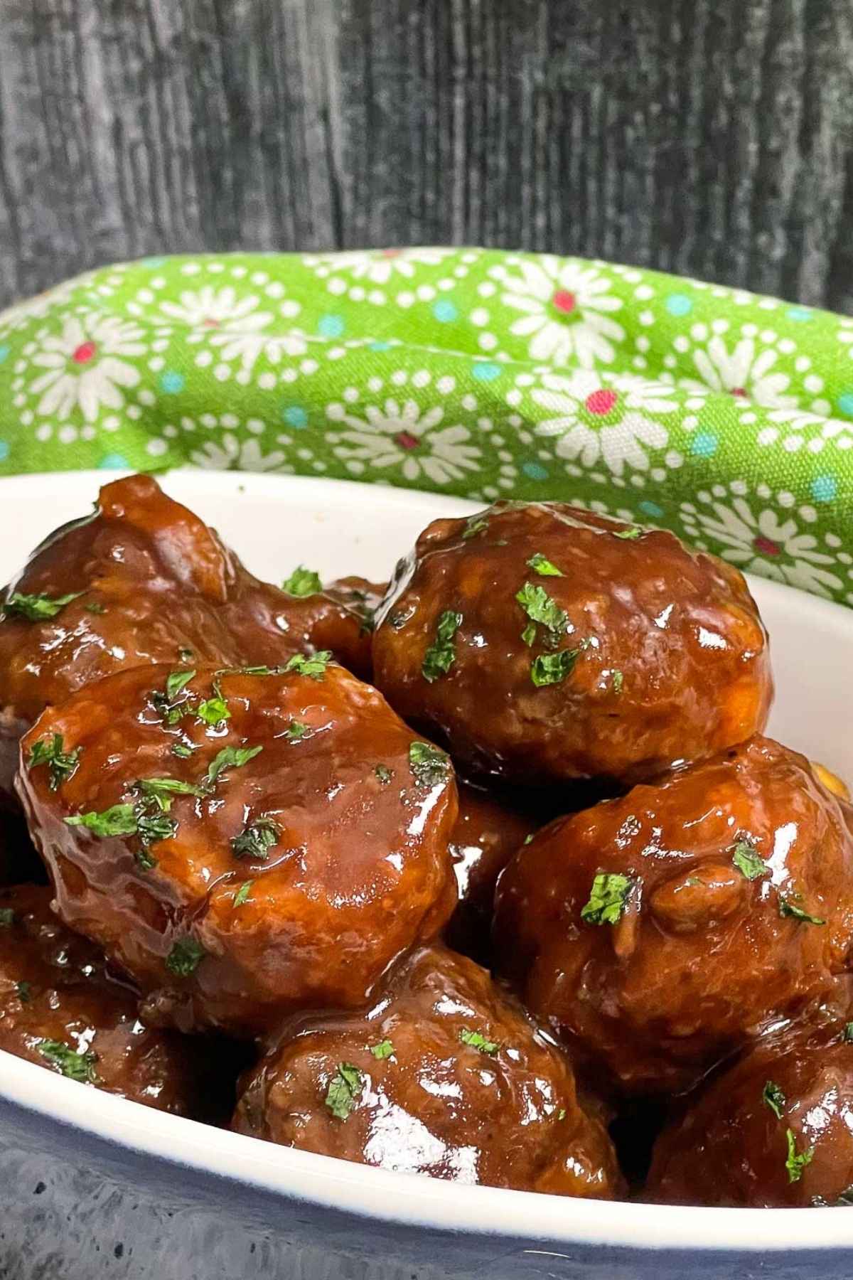 Close up of grape jelly meatballs in a white and blue oval dish, coated in a shiny sauce and sprinkled with fresh parsley, with a green floral napkin in the background.