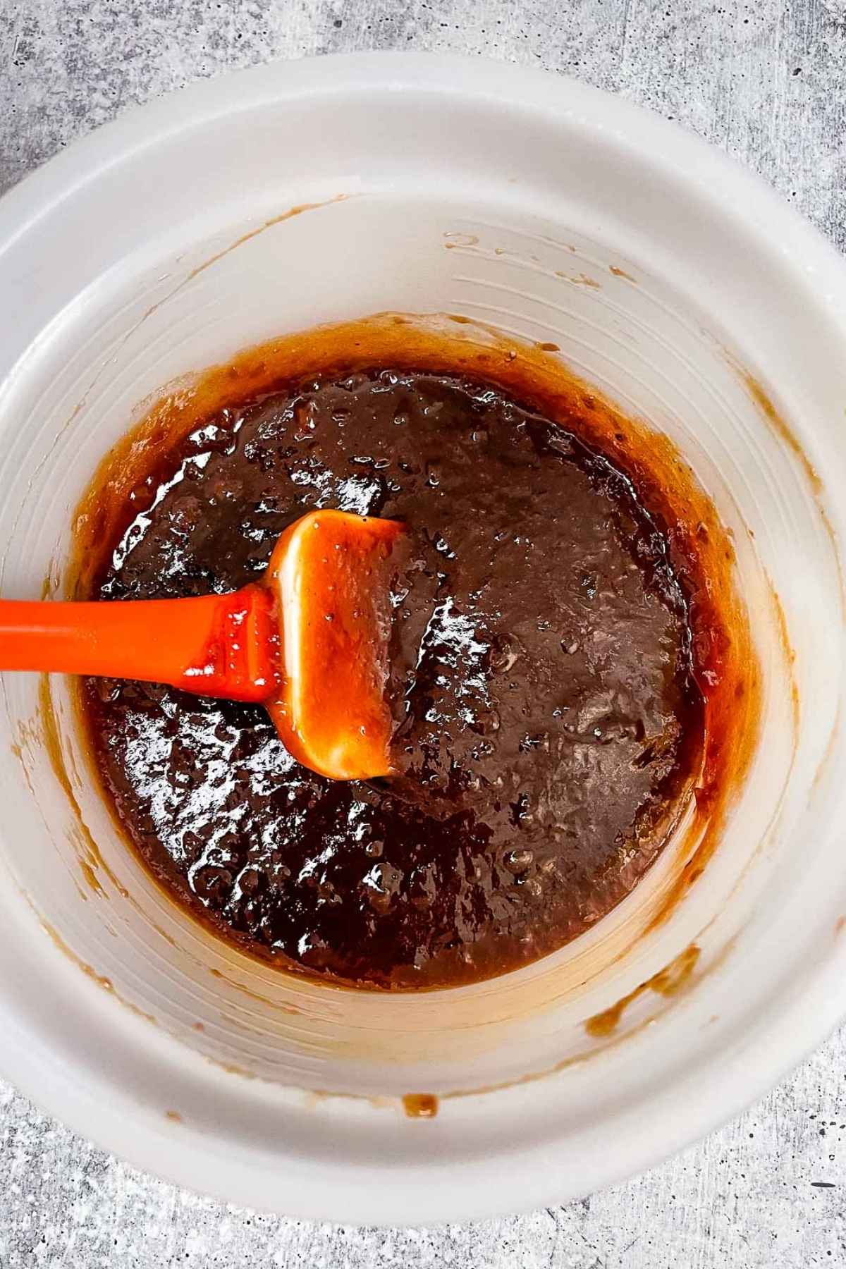 Mixing bowl with thick grape jellyBBQ sauce and an orange spatula, showing texture and consistency of the sauce.