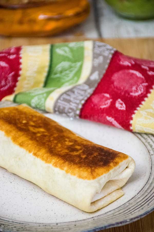 A grilled stuffed burrito with a golden brown crispy exterior sits on a speckled ceramic plate, with a colorful cloth napkin in the background.