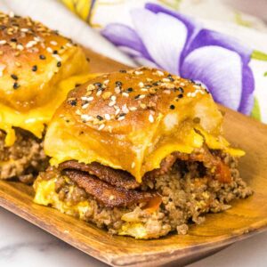 Two cheesy ground beef sliders on a wooden plate, filled with ground beef, melted cheese, and bacon, topped with golden everything bagel seasoned buns.