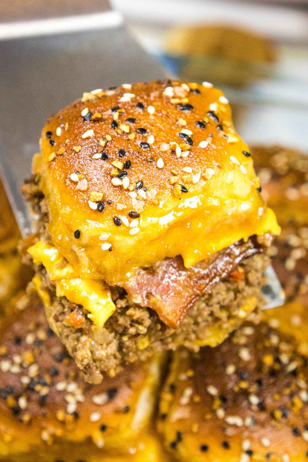 Close up of a single baked slider being lifted with a spatula, showing melted chese, crispy bacon, and seasoned ground beef filling inside golden, sesame topped buns.