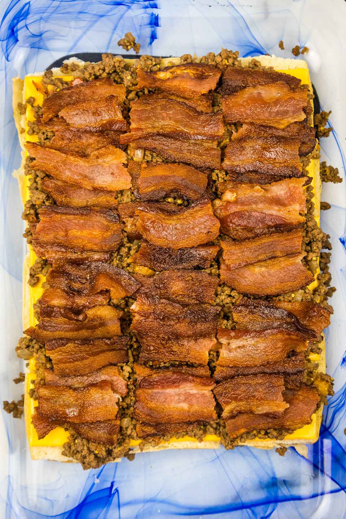Overhead view of ground beef slider filling in a baking dish, showing a layer of crispy cooked bacon peices on top of seasoned ground beef and cheese slices.