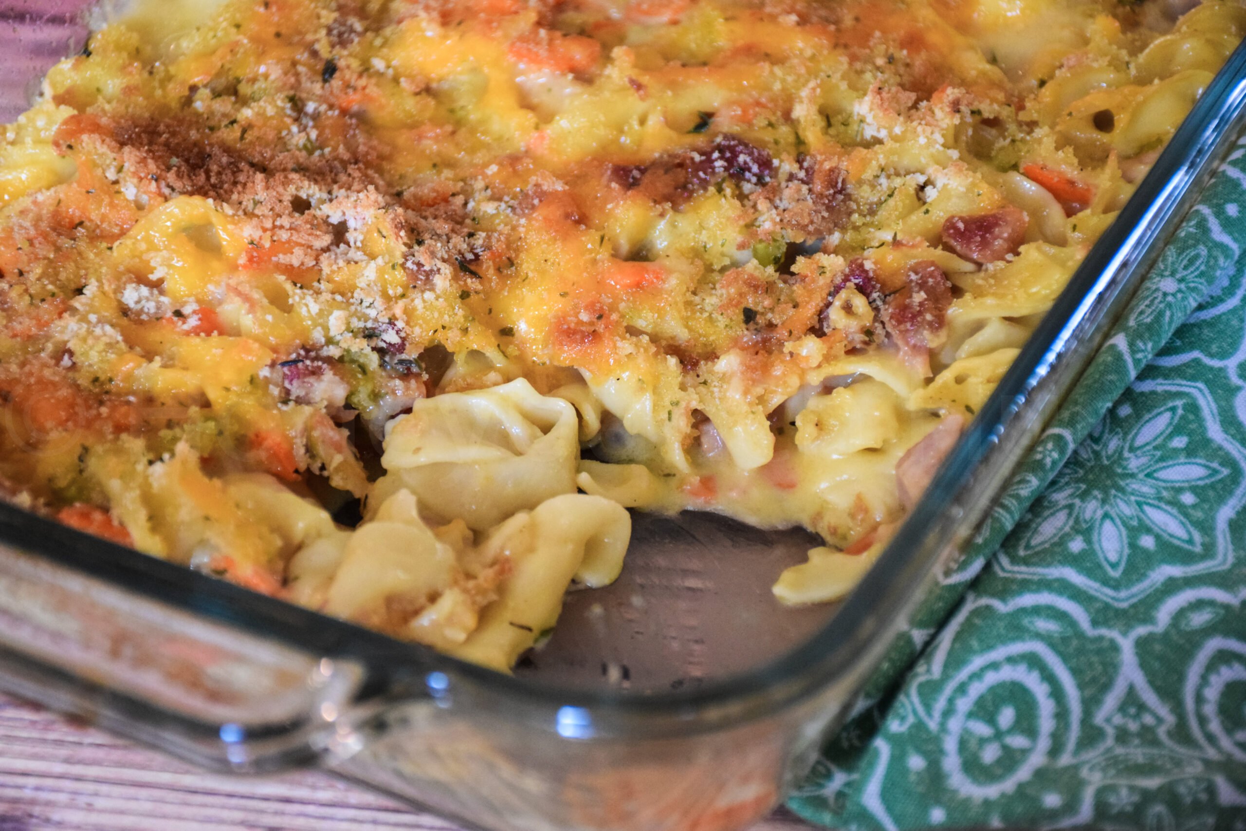 A glass baking dish of ham and noodle casserole with a golden cheesy breadcrumb topping, showing a serving removed to reveal the creamy pasta, diced ham, and vegetables inside.
