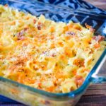 A glass baking dish filled with baked ham and noodle casserole, featuring wide egg noodles, chunks of ham, melted cheddar cheese, and a golden breadcrumb topping.