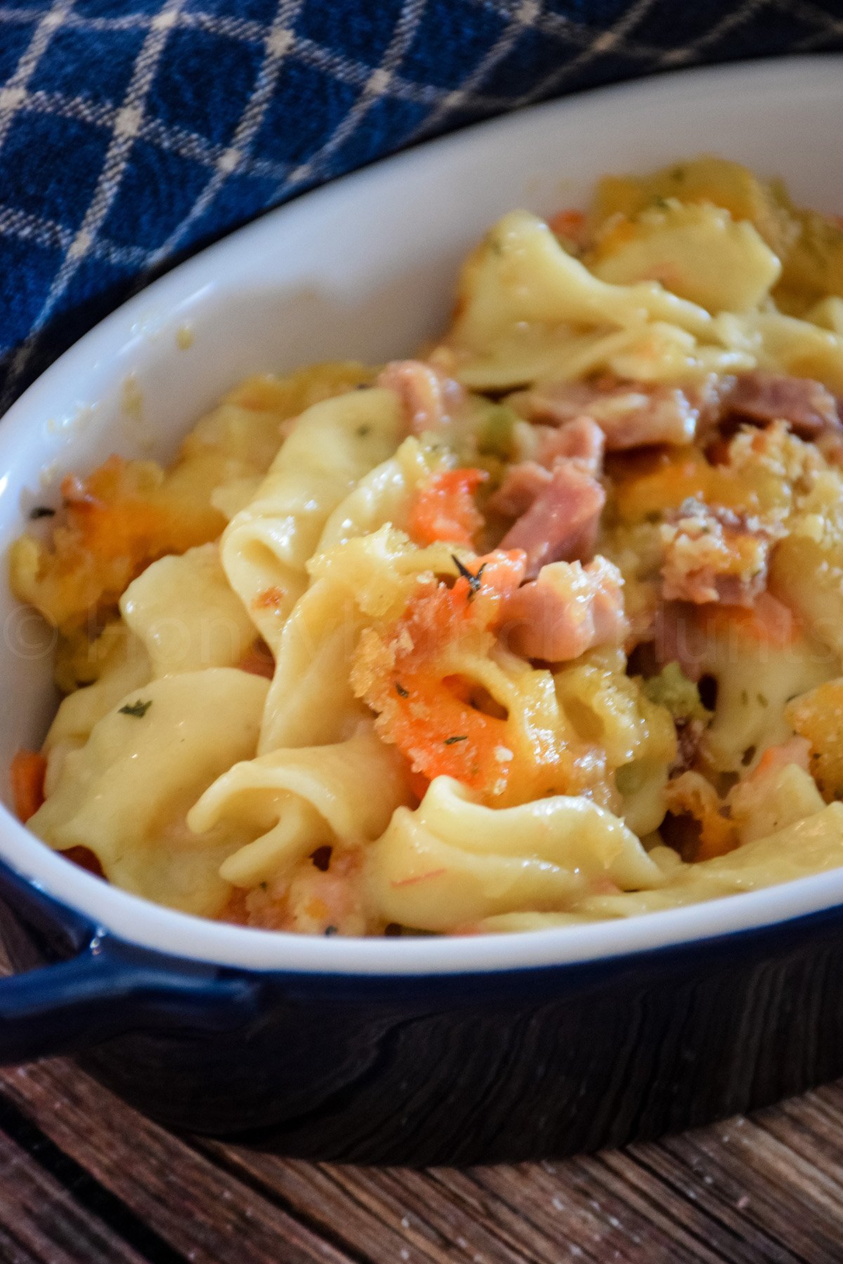 A serving of baked ham and noodle casserole in a white dish, featuring cheesy egg noodles, bits of ham, carrots, and golden breadcrumb topping.