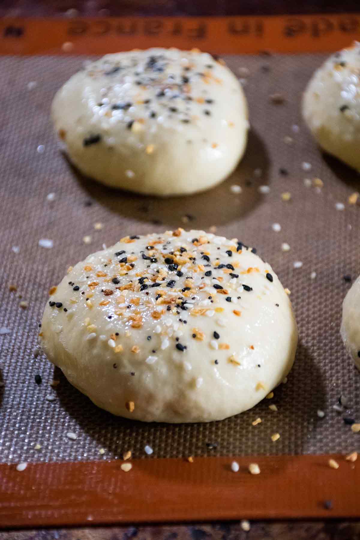 Raw homemade hamburger bun dough on baking mat, sprinkled with everything seasoning and ready to bake.