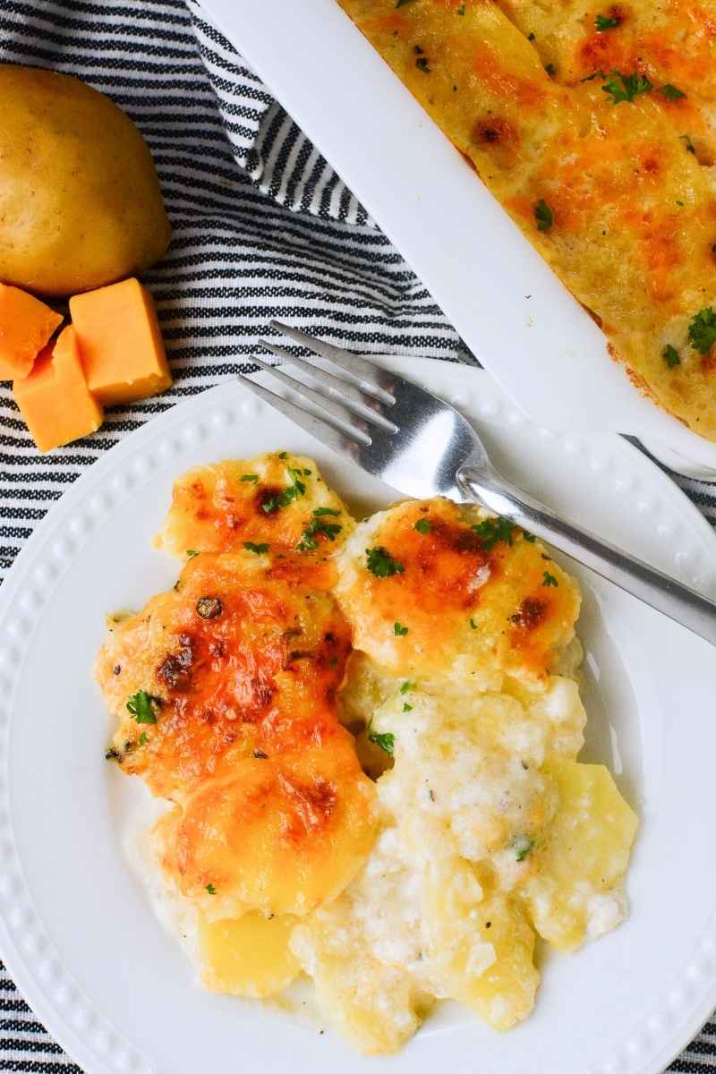 A white plate with a hearty serving of scalloped potatoes topped with parsley, set beside a casserole dish and with cheddar cheese cubes and a whole potato in the background.