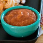 Close-up of a turquoise bowl filled with thick, reddish-brown homemade cocktail sauce, served on a black plate with shrimp in the background.