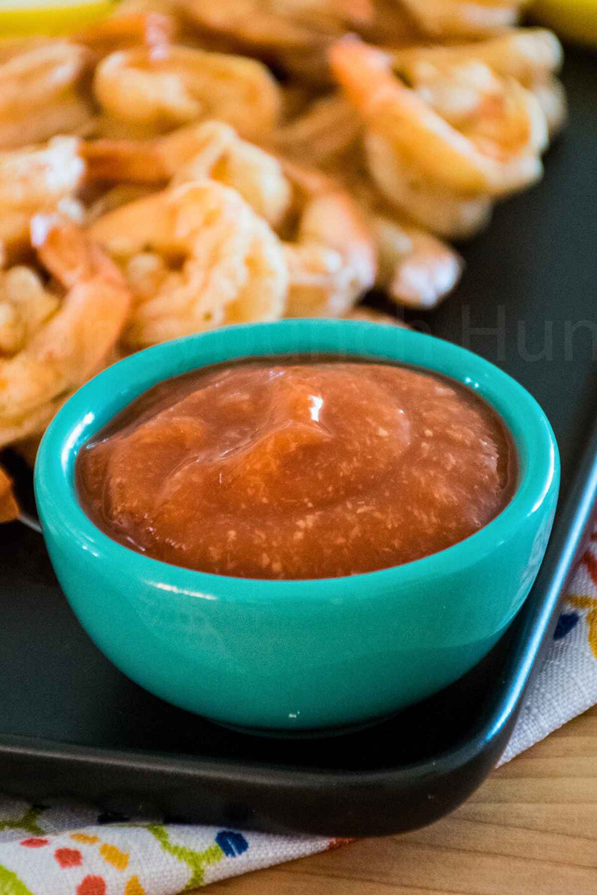 A bowl of cocktail sauce with shrimp in the background on a black plate..
