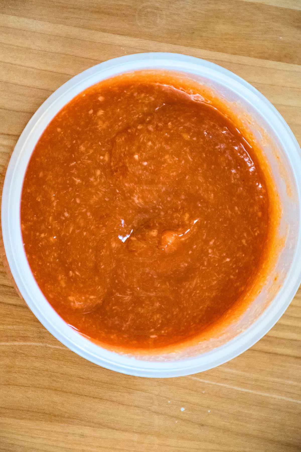 Overhead view of sauce in a white plastic mixing bowl showing a thick, textured reddish sauce on a wooden surface.