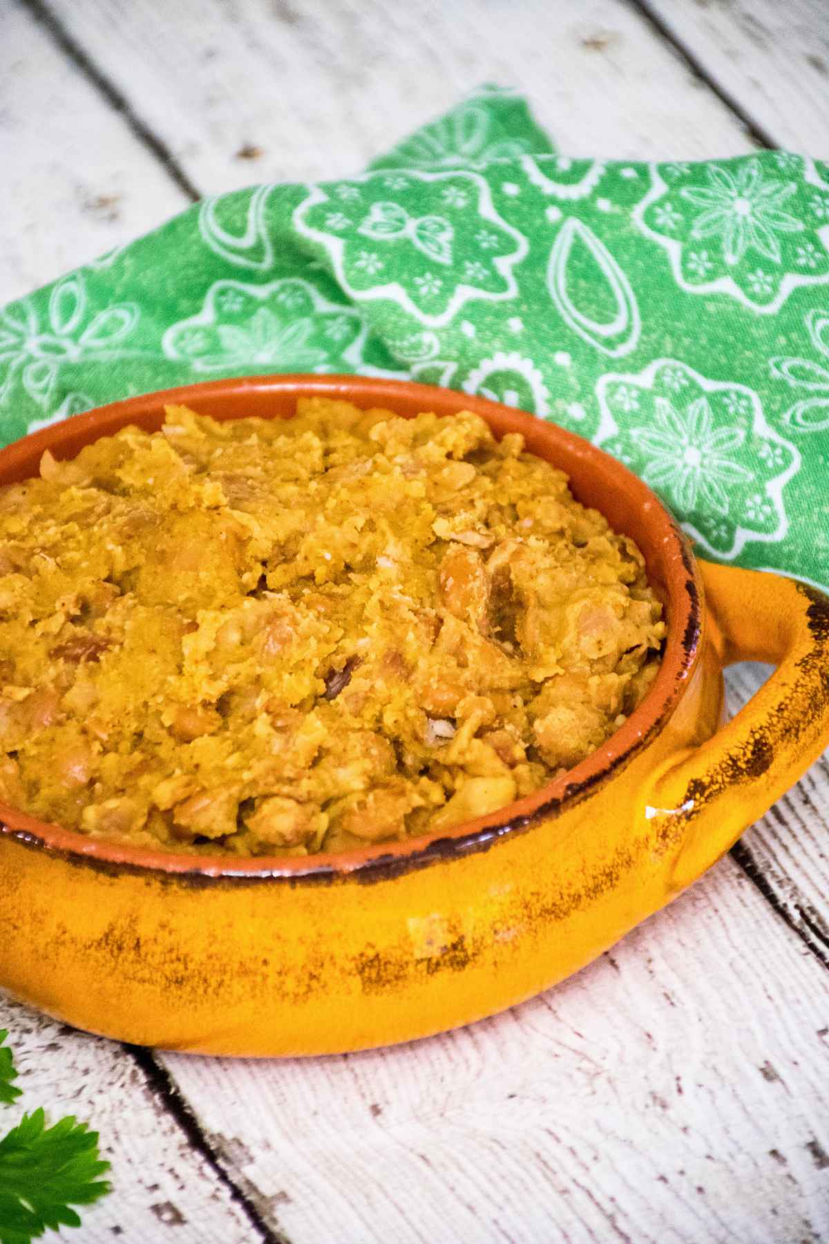 Angled view of a rustic yellow ceramic bowl filled with homemade refried beans, set on a white wooden surface with a green patterned cloth in the background.
