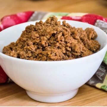 A white bowl filled with cooked, seasoned ground taco meat, set on a wooden surface with a colorful patterned towel in the background.
