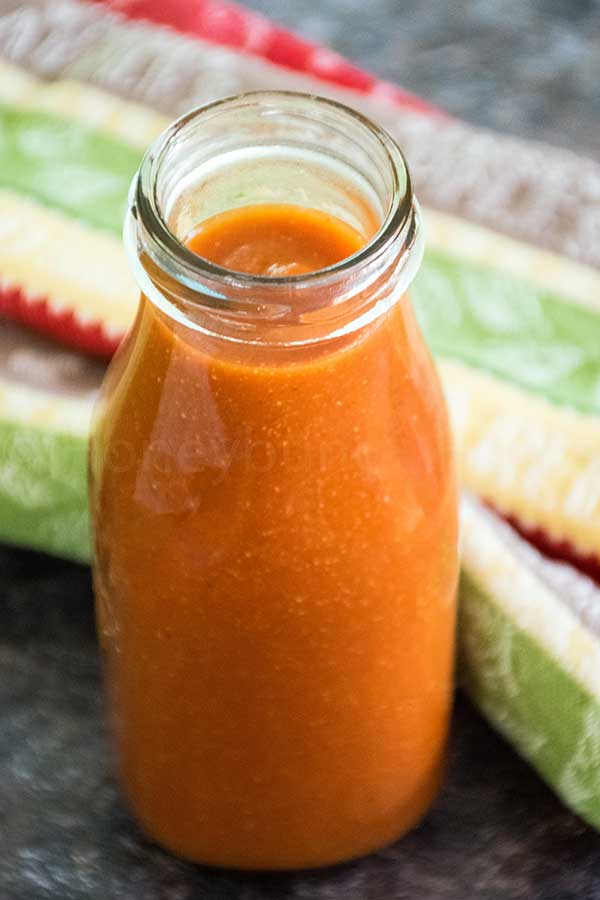 A glass bottle filled with vibrant homemade taco sauce, set on a dark countertop with a colorful striped towel in the background.