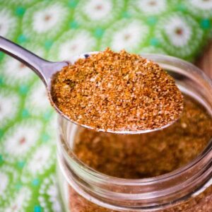 A spoonful of finely blended homemade taco seasoning held above a jar with a background of a green cloth patterend with white daisies.