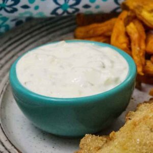 A small turquoise bowl filled with creamy homemade tartar sauce, served alongside golden sweet potato fries and a breaded fish fillet on a speckled plate.