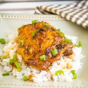 Close up of a honey chipotle glazed chicken thigh on a bed of white rice, garnished with chopped scallions, served on a square cream colored plate.