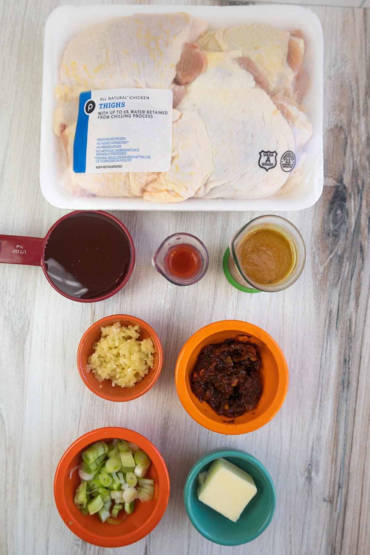 Overhead view of raw chicken thighs in a package with ingredients for honey chipotle sauce, including chopped garlic, green onions, chipotle peppers, butter, honey, hot sauce, and soy sauce in small bowls and measuring cups.