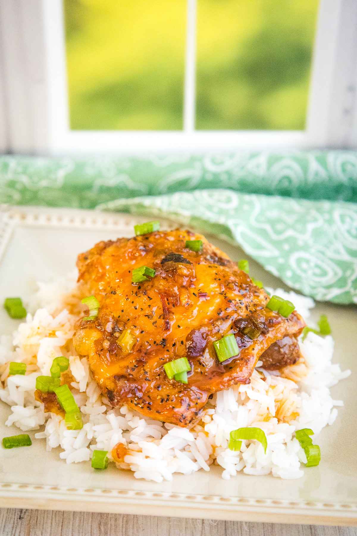Close up of a honey chipotle glazed chicken thigh on a bed of white rice, garnished with chopped scallions, served on a square cream colored plate.