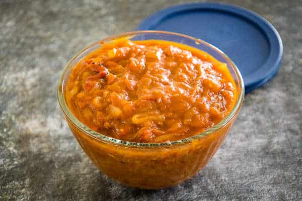 Side angle of a glass bowl filled with vibrant orange-red spicy tomato chutney, set on a gray surface with a blue lid in the background.