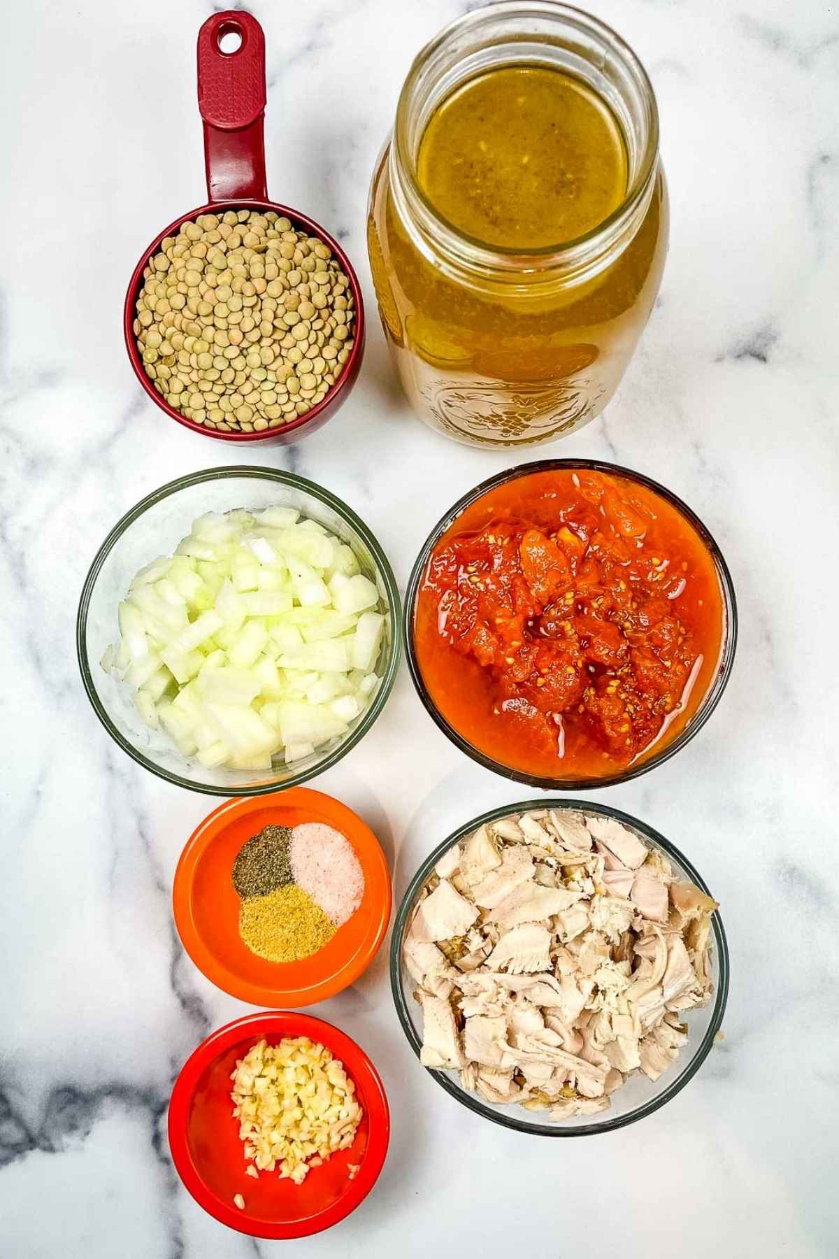 Overhead view of measured ingredients on a white marbled surface, including dry lentils, diced onions, canned tomatoes, chopped cooked turkey, minced garlic, seasoning mix, and a jar of broth.