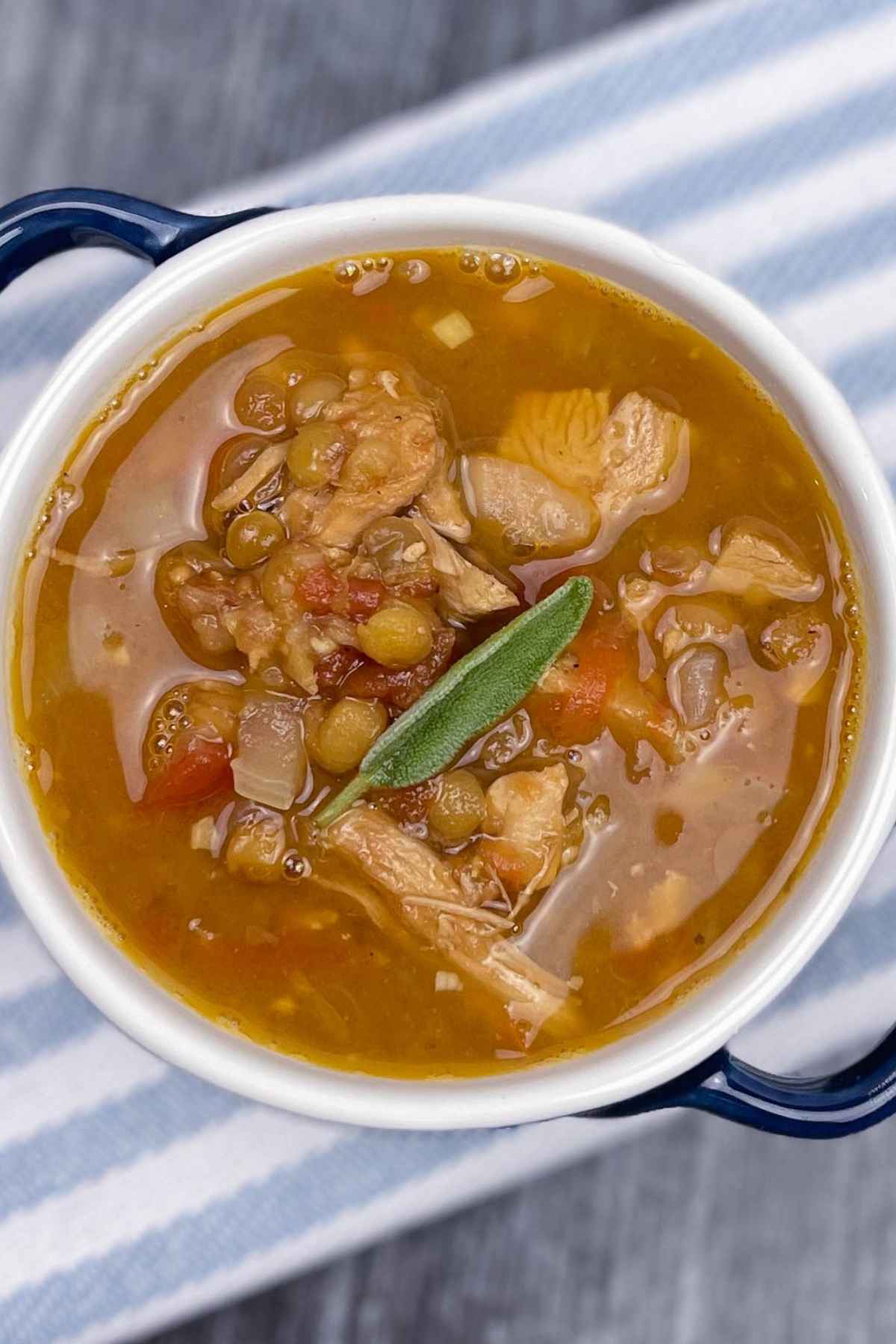 Overhead view of a soup crock filled with turkey lentil soup, garnished with a sage leaf, sitting on a striped cloth over a dark wooden surface.