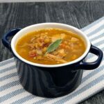 A small navy blue soup crock filled with turkey lentil soup, garnished with a sage leaf, sitting on a striped cloth over a dark wooden surface.