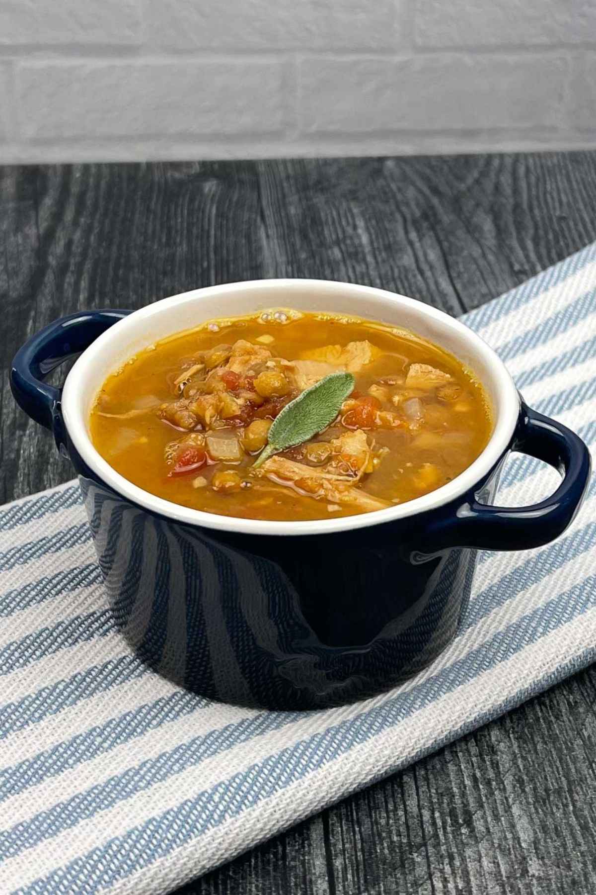 A small navy blue soup crock filled with turkey lentil soup, garnished with a sage leaf, sitting on a striped cloth over a dark wooden surface.