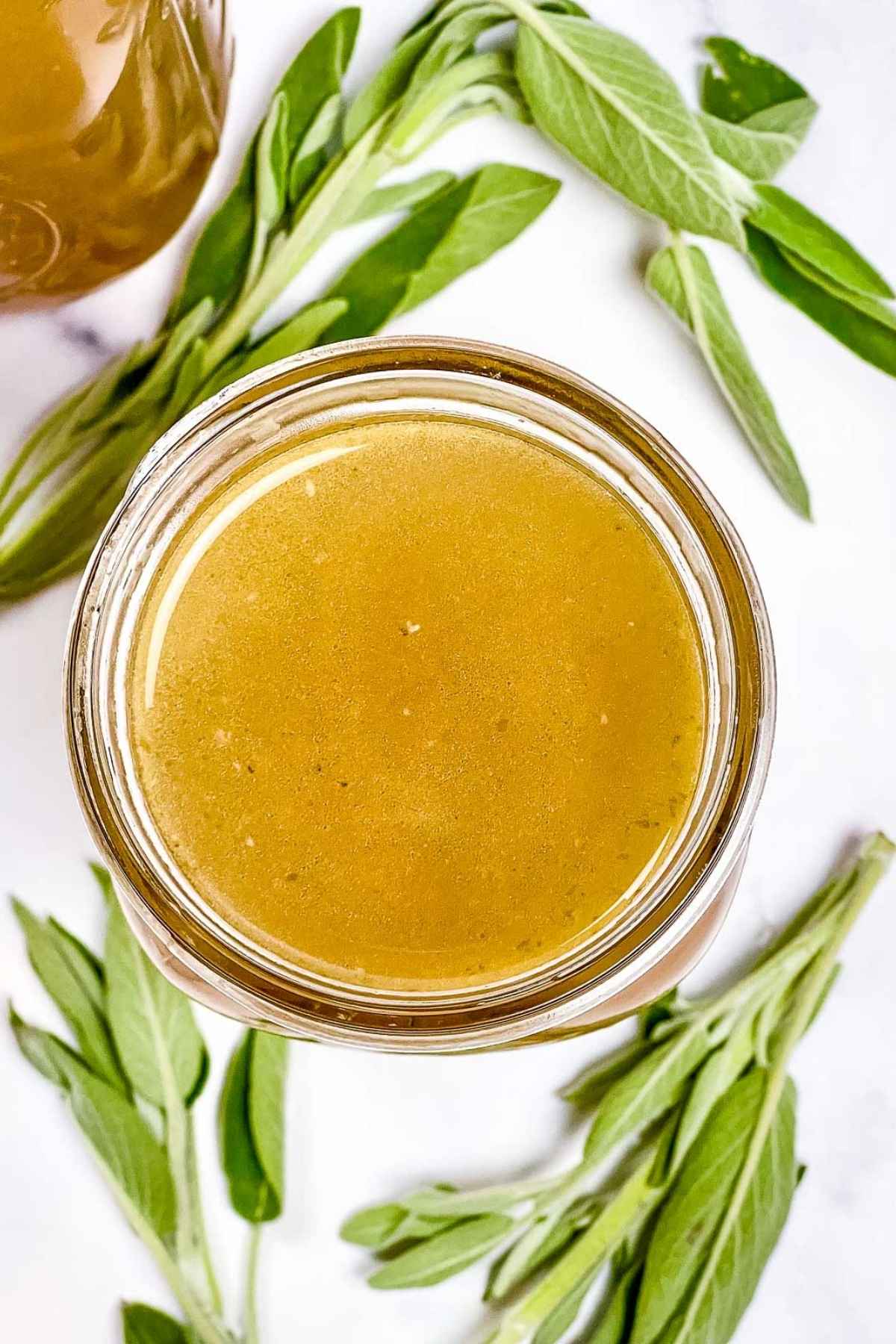 Overhead shot of a glass jar filled with homemade turkey stock, with sprigs of fresh sage scattered around on a white background.