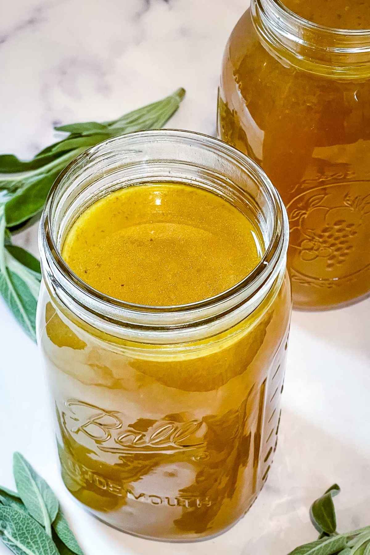 Close up side view of a Ball mason jar filled with golden turkey stock, with another jar in the background and sage leaves nearby.