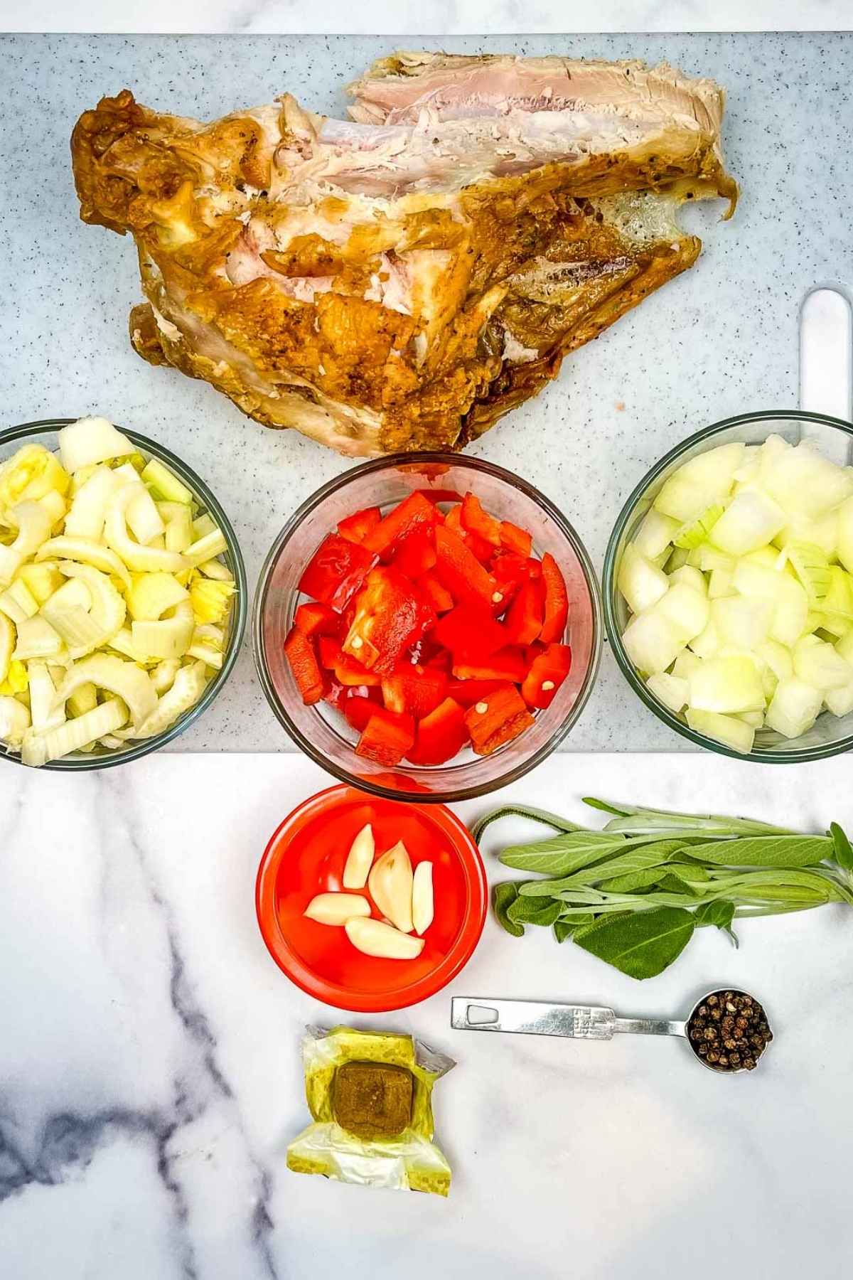 Assorted ingredients laid out on a marble counter, including a turkey carcass, chopped onions, celery, red peppers, garlic, sage, bouillon cube, and whole black peppercorns in a measuring spoon.