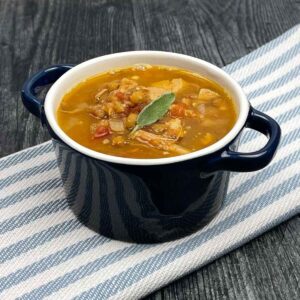A small navy blue soup crock filled with turkey lentil soup, garnished with a sage leaf, sitting on a striped cloth over a dark wooden surface.