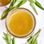 Overhead shot of a glass jar filled with homemade turkey stock, with sprigs of fresh sage scattered around on a white background.