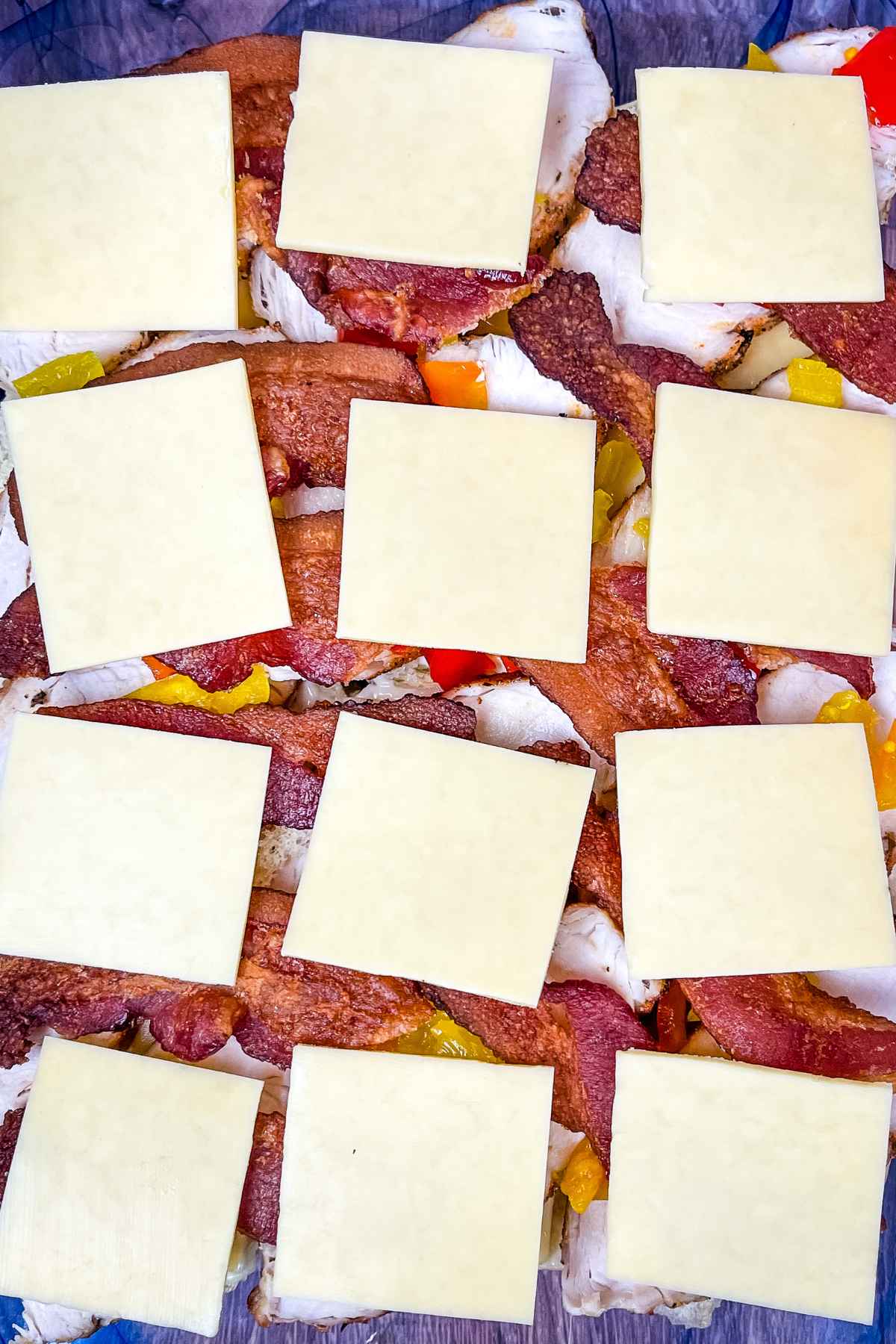 Kentucky Hot Brown Sliders in a glass baking dish topped with turkey peppers, bacon, and cheese before placing the slider roll tops on.