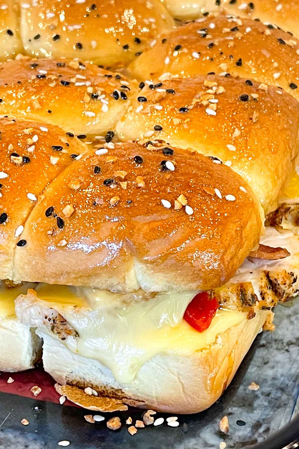 Close up of golden brown baked sliders topped with everything bagel seasoning, filled with melted cheese, sliced turkey, and red peppers on a dark tray.