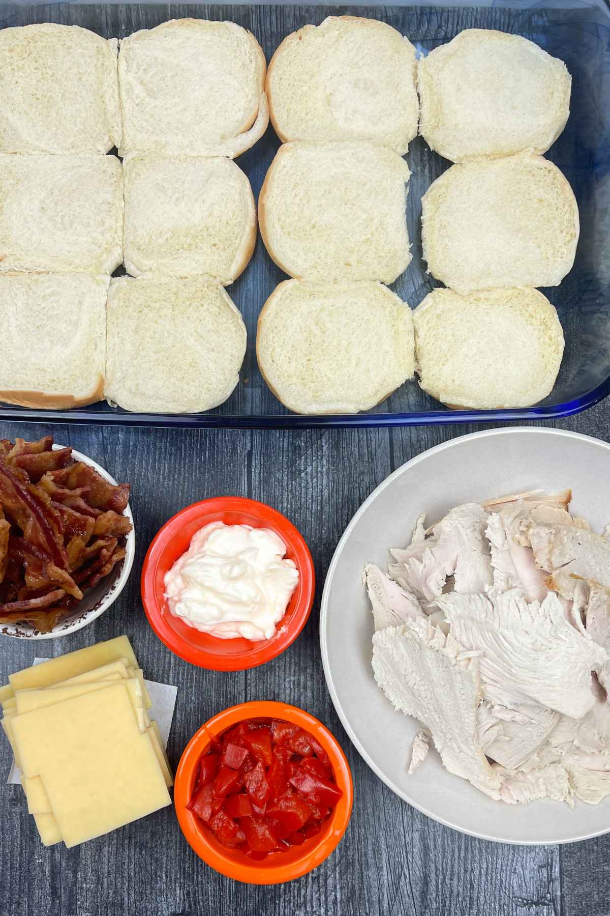 Overhead view of ingredients for Kentucky Hot Brown Sliders, including halved slider buns in a baking dish, cooked bacon strips, sliced turkey, Swiss cheese, chopped red peppers, and a bowl of mayonnaise on a dark wooden surface.