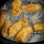 Golden grown breaded gluten Free Fish Sticks sizzling in hot oil inside a black skillet during the frying process.
