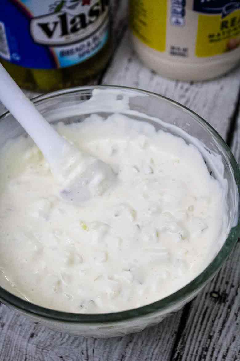 A glass mixing bowl with freshly prepared sauce with visible bits of onion and pickle, with a spatula resting inside and ingredient jars nearby.