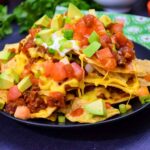 A heaping plate of loaded nachos piled high with melted cheese, ground meat, diced tomatoes, avocado chunks, salsa, sour cream, and sliced green onions, served on a black plate.