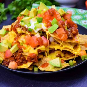 A heaping plate of loaded nachos piled high with melted cheese, ground meat, diced tomatoes, avocado chunks, salsa, sour cream, and sliced green onions, served on a black plate.