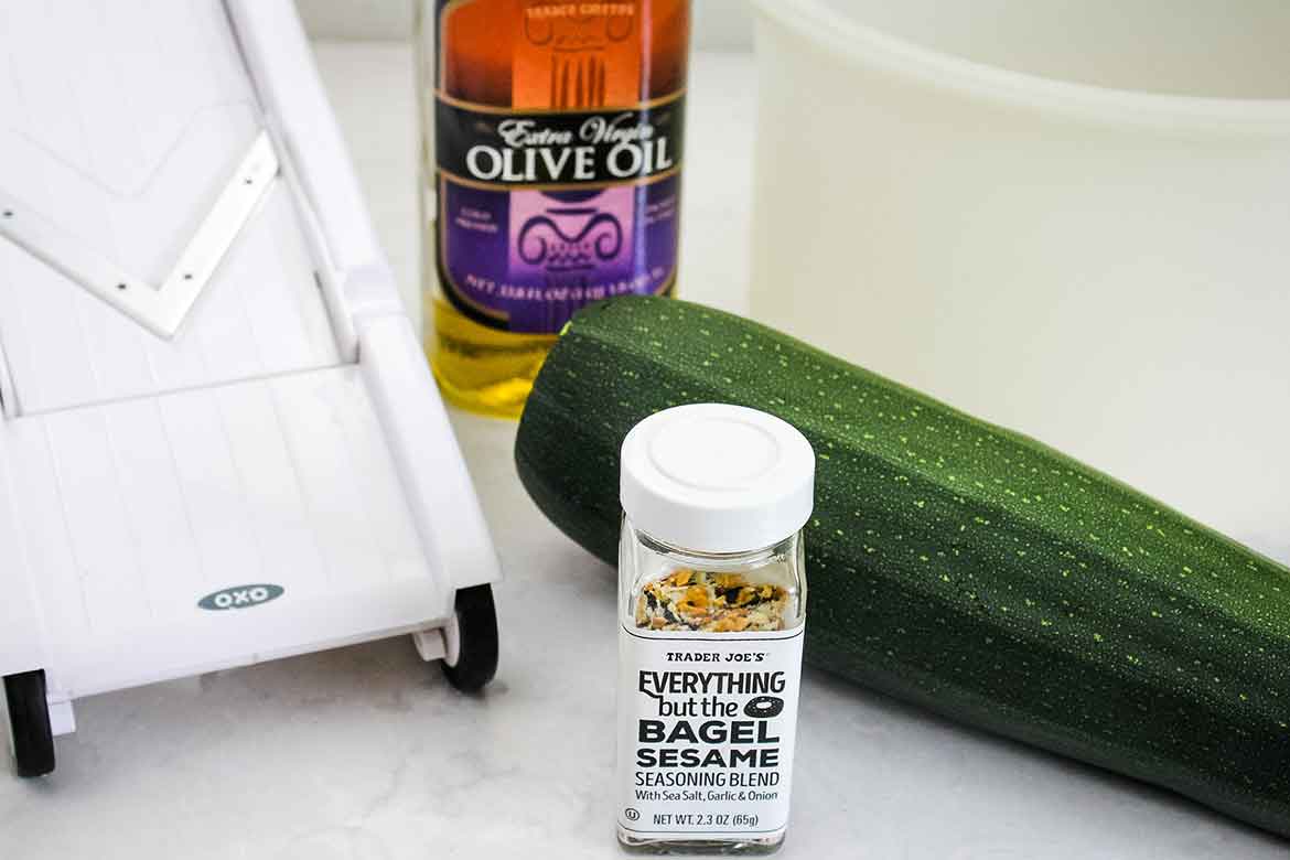 Ingredients for zucchini chips: whole zucchini, a bottle of olive oil, everything bagel seasoning, and a white mandoline slicer.